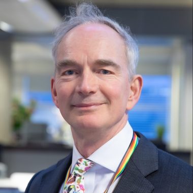 A professional-looking individual wearing a suit and a colorful tie with a rainbow lanyard around the neck.
