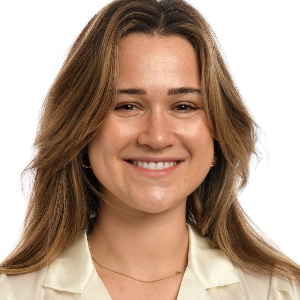 A person with long brown hair smiles, wearing a cream-colored blouse against a plain white background.