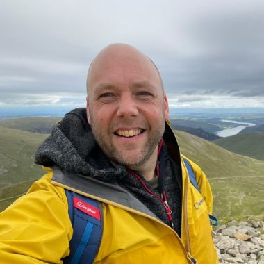 A smiling bald man in a yellow jacket takes a selfie on a mountain summit with a lake and hills in the background.