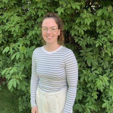 A person with glasses stands smiling in front of a leafy green background, wearing a white and gray striped shirt and light-colored pants.