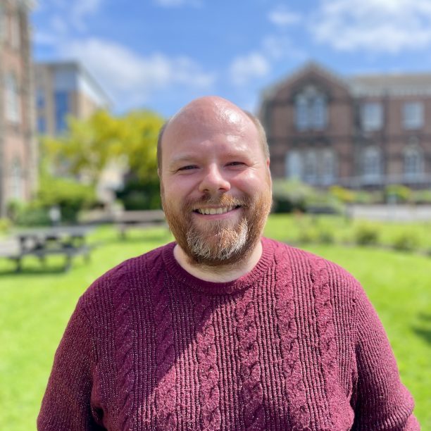 A man with a beard and shaved head smiles while standing outdoors under a clear sky, with green grass and a building in the background.