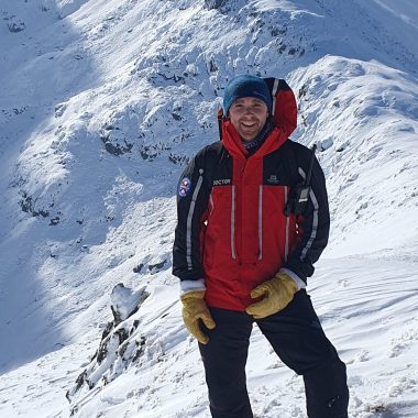 A person in a red and black winter jacket, blue hat, and yellow gloves stands on a snowy mountain with snow-covered peaks in the background.