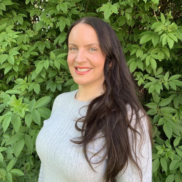 A woman with long dark hair, wearing a gray sweater, smiling and standing in front of a leafy green background.