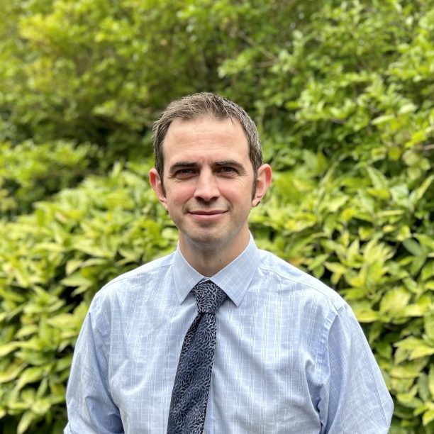 A person in a light blue shirt and dark tie stands in front of lush green foliage.