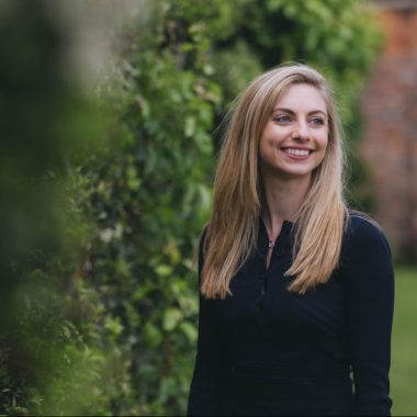 A woman with long blonde hair, wearing a black top, stands outdoors smiling. She is surrounded by greenery and trees.