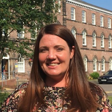 A woman with long brown hair and a floral top smiles outdoors in front of a historic brick building with arched windows. A tree and parked car are visible in the background.