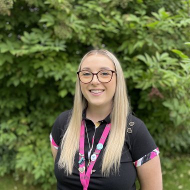 A woman with long blonde hair and glasses stands outdoors, wearing a black shirt adorned with pins and a pink lanyard, smiling in front of greenery.