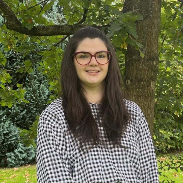 A woman with long dark hair and glasses stands in front of a tree on a grassy area, wearing a black and white checkered shirt and black pants.