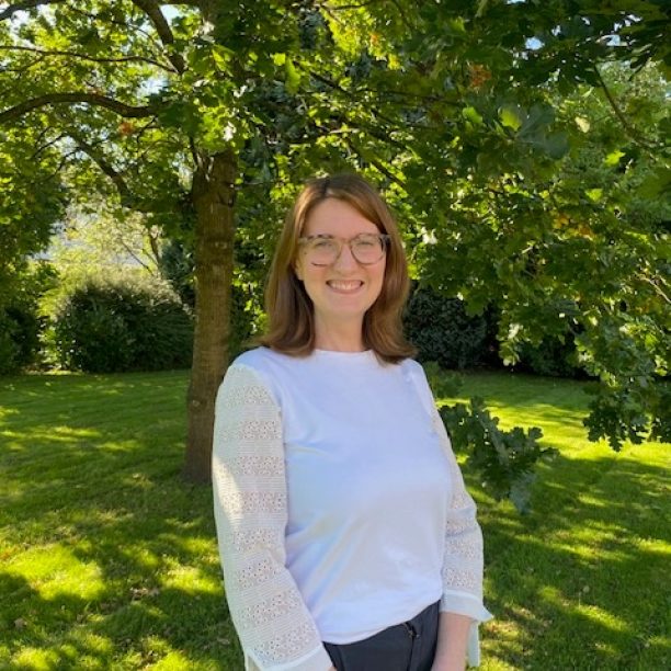 A person with shoulder-length brown hair and glasses stands outdoors in front of a tree, smiling, wearing a white blouse with lace sleeves and dark pants.