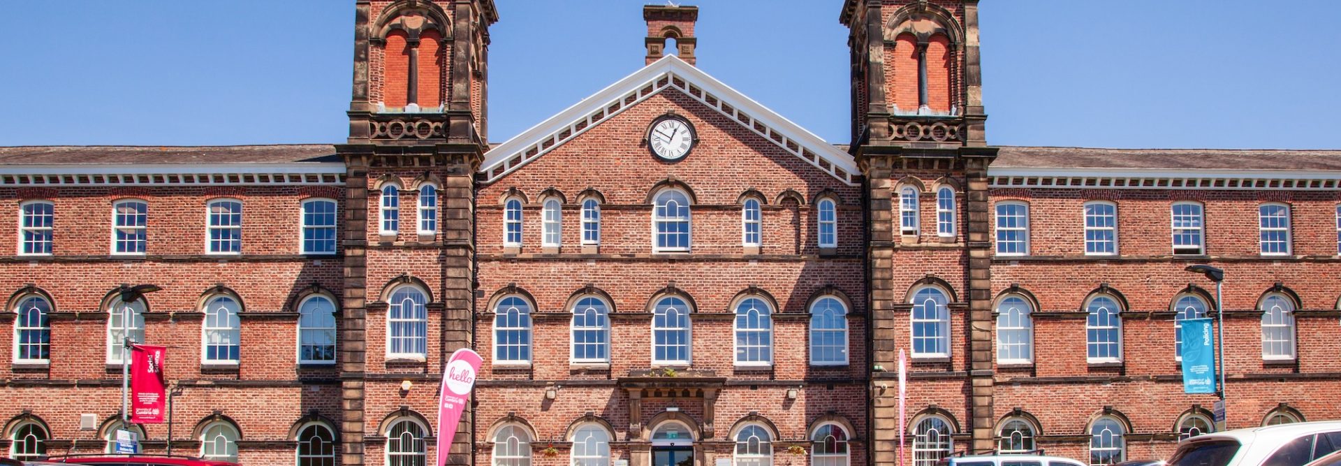 Red brick building with distinctive twin towers and arched windows, functioning as a modern facility with a parking lot in the foreground.