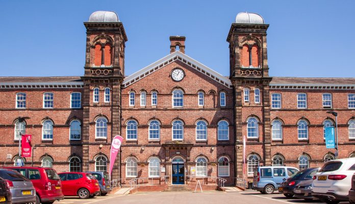 Red brick building with distinctive twin towers and arched windows, functioning as a modern facility with a parking lot in the foreground.