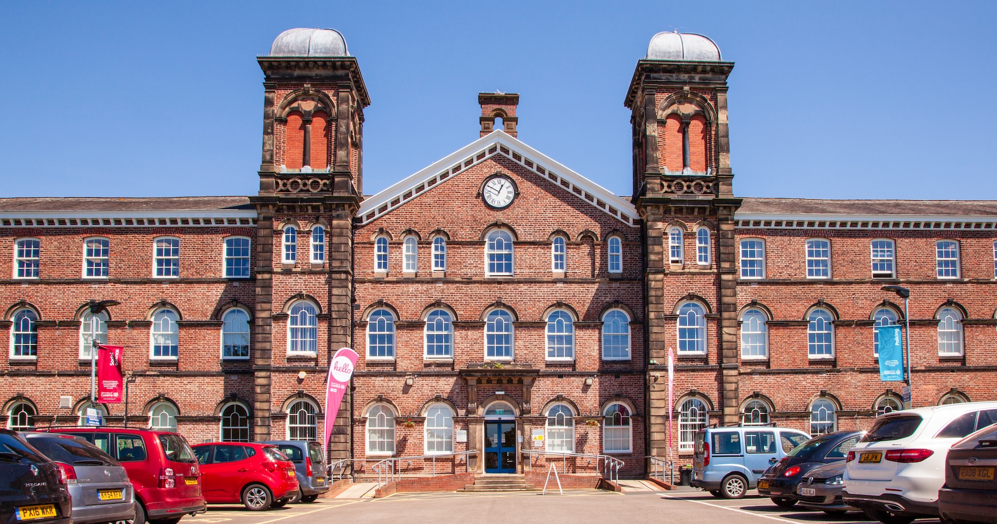 Red brick building with distinctive twin towers and arched windows, functioning as a modern facility with a parking lot in the foreground.