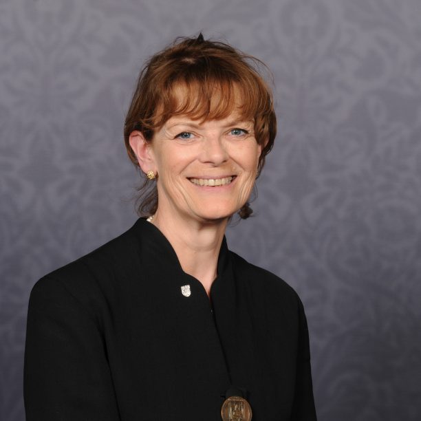 A person with short hair, dressed in a black outfit and a pin symbolizing medical school pride, smiles against a patterned background.