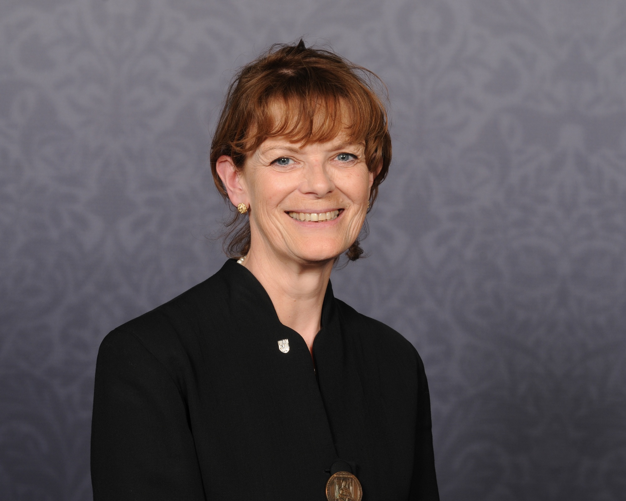 A person with short hair, dressed in a black outfit and a pin symbolizing medical school pride, smiles against a patterned background.