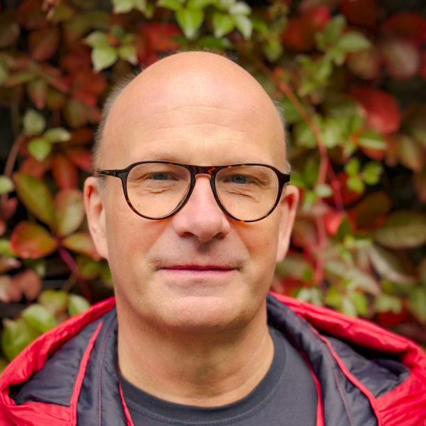 A person with glasses and a red jacket stands in front of a background of red and green leaves.