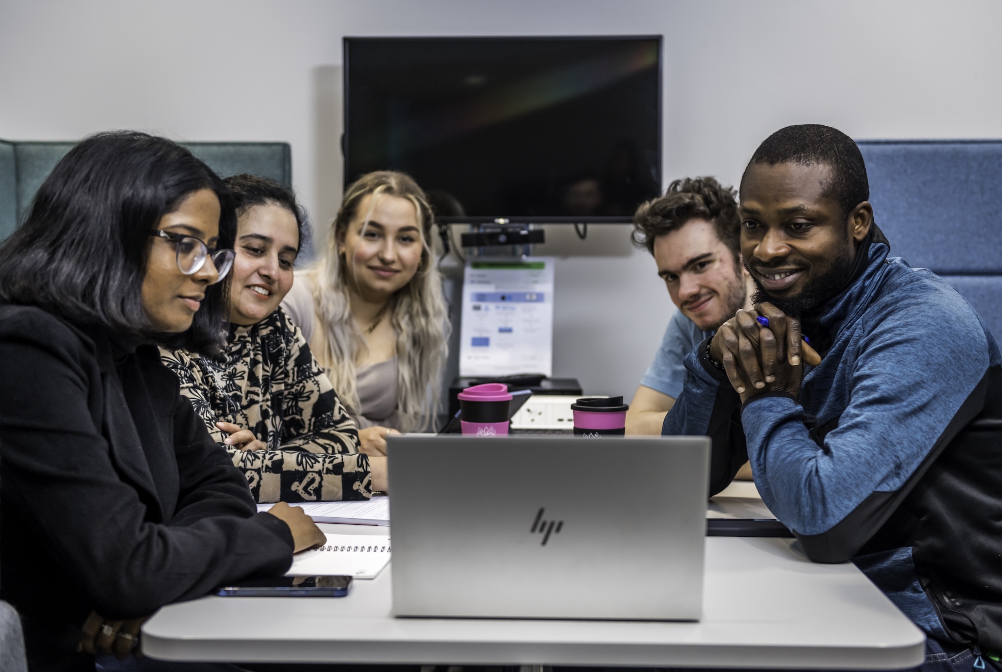 Five medical school students sit at a table, focused on a laptop screen, with a monitor and notepad in the background.